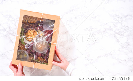 Flat lay. Hands are meticulously adding fresh red grapes to a bowl, complementing a beautifully arranged charcuterie box brimming with a variety of cheeses, olives, and cured meats, set against a 123355156