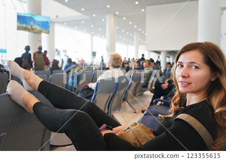 Young woman sitting at airport hall, looking tired after waiting couple of hours for connecting flight in early morning, legs on seat in front of of her, more blurred passengers in background Young woman sitting at airport hall, looking tired after waiting couple of hours for connecting flight in early morning, legs on seat in front of of her, more blurred passengers in background 123355615