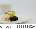 Side view of mini desserts lying on a plate, behind them is a white mug with a hot drink on a white background with space for text. The concept of mini desserts, sweets, confectionery. High quality 123355620