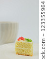 A cake consisting of an airy sponge cake and white cream, decorated with a mastic flower on top, stands on a white saucer in close-up against the background of a white mug with a hot drink. The 123355964