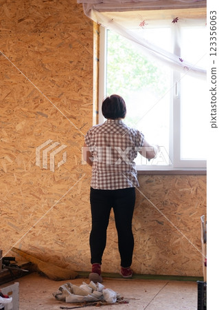 Woman installs a bright, white window in a wood paneled room with curtains. She is wearing black pants, a plaid shirt and red shoes. 123356063
