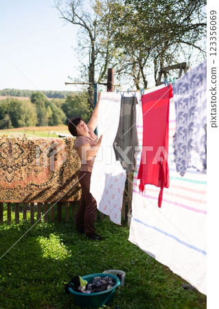 Woman hanging laundry on a clothesline in her yard on a sunny day, with a rustic rug hanging on the fence behind her 123356069