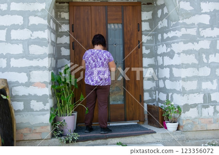 Woman at Front Door: Ready to go inside. She's facing a wooden door with brickwork and potted plants adding a touch of nature. Woman at Front Door: Ready to go inside. She's facing a wooden door with brickwork and potted plants adding a touch of nature. 123356072
