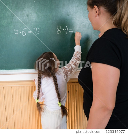 Young student calculates multiplication problem on chalkboard with teacher observing in classroom, focusing on education and development. Young student calculates multiplication problem on chalkboard with teacher observing in classroom, focusing on education and development. 123356077