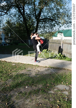 A woman in black leggings is holding a child with a red backpack in a grassy yard by a weathered fence under a large tree. 123356087