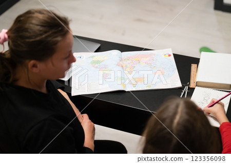 A mother and daughter engage in homeschooling, looking at a world map while the child draws in a notebook, fostering education at home. 123356098