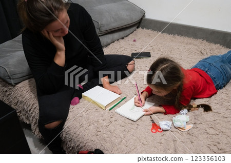 A young girl is drawing on a notebook while laying on a carpet next to a woman who is sitting with her head in her hand. 123356103