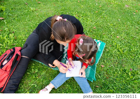 A woman and a child are drawing together outdoors on a blanket. They are enjoying a sunny day on the green grass. 123356111