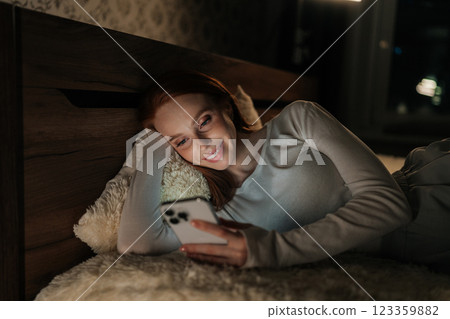 Charming young woman lying in bed at night, face illuminated by soft glow of smartphone, smiling scrolling social media content and enjoying moment of connection. Concept of addiction and insomnia. 123359882
