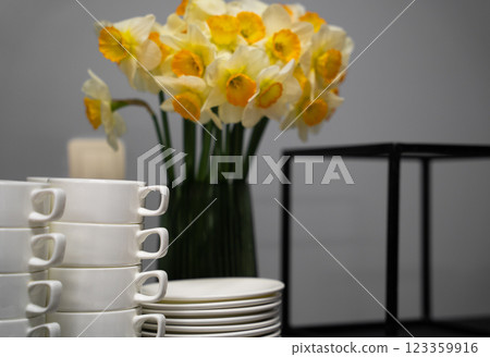 Many clean white cups and plates on the table near the coffee maker and vase with daffodils Many clean white cups and plates on the table near the coffee maker and vase with daffodils 123359916