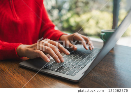 Close-up of hands typing on a laptop in a cafe, with natural light and greenery outside. 123362876