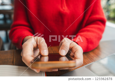 Hands holding a smartphone, wearing a red sweater, at a wooden table, capturing a moment of modern technology use. Hands holding a smartphone, wearing a red sweater, at a wooden table, capturing a moment of modern technology use. 123362878