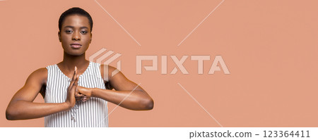 African American young woman wearing a striped tank top practices yoga with her hands in front of her chest, in a meditative pose. She is in front of a peach-colored background. 123364411