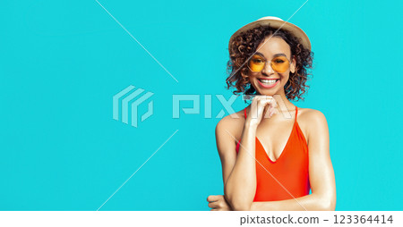 This image features a young woman wearing a red swimsuit and a straw hat. She is smiling and looking at the camera, with her hand resting on her chin. She is standing against a solid blue background. This image features a young woman wearing a red swimsuit and a straw hat. She is smiling and looking at the camera, with her hand resting on her chin. She is standing against a solid blue background. 123364414
