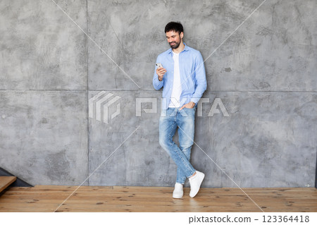 A man in a blue button-down shirt and jeans stands with one leg crossed against a concrete wall, looking at his smartphone with a slight smile. The man has a beard and short dark hair, copy space A man in a blue button-down shirt and jeans stands with one leg crossed against a concrete wall, looking at his smartphone with a slight smile. The man has a beard and short dark hair, copy space 123364418