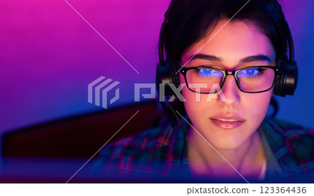 A woman wearing glasses and headphones is looking intently at a computer screen. The room is lit in a purple and blue color scheme, creating a vibrant and modern atmosphere A woman wearing glasses and headphones is looking intently at a computer screen. The room is lit in a purple and blue color scheme, creating a vibrant and modern atmosphere 123364436
