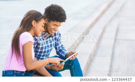 African American teen couple, sitting on steps outside, smiles as they look at a tablet. The girl is wearing a pink shirt and jeans, while the boy is wearing a plaid shirt and blue jeans. African American teen couple, sitting on steps outside, smiles as they look at a tablet. The girl is wearing a pink shirt and jeans, while the boy is wearing a plaid shirt and blue jeans. 123364437