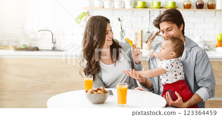 A happy family enjoys a meal together in their kitchen. The father holds their baby, who is reaching out to the mother who sits across the table, holding a yellow toy in her hand, copy space A happy family enjoys a meal together in their kitchen. The father holds their baby, who is reaching out to the mother who sits across the table, holding a yellow toy in her hand, copy space 123364459
