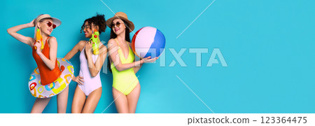 This image shows three women in swimsuits posing against a blue background. They are holding beach toys, including a beach ball and water guns. The women are smiling and appear to be having fun. This image shows three women in swimsuits posing against a blue background. They are holding beach toys, including a beach ball and water guns. The women are smiling and appear to be having fun. 123364475