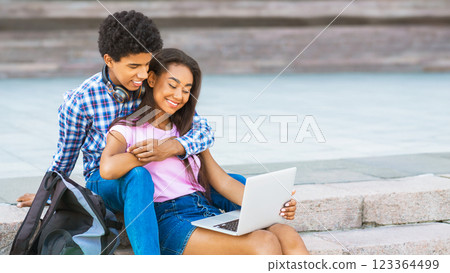 A teen couple is sitting on stone steps outside, the girl is using a laptop and guy has his arm around her. They are both smiling. The image captures a moment of intimacy and connection, copy space A teen couple is sitting on stone steps outside, the girl is using a laptop and guy has his arm around her. They are both smiling. The image captures a moment of intimacy and connection, copy space 123364499
