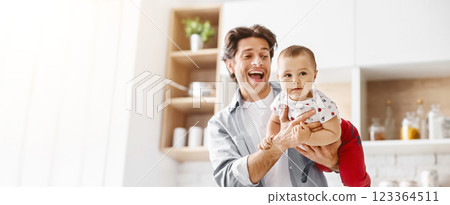 The image shows a happy father holding his baby in the kitchen. The father is smiling and looking at the camera while the baby is looking at something off-camera, copy space 123364511