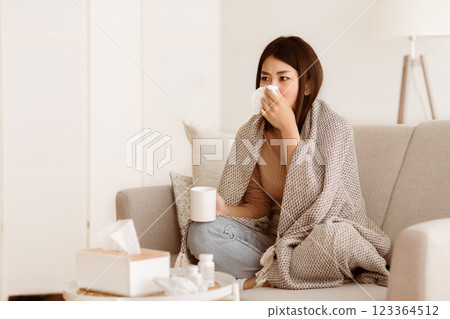 A woman sits on a couch at home, wrapped in a blanket, blowing her nose while holding a cup. She looks unwell. A woman sits on a couch at home, wrapped in a blanket, blowing her nose while holding a cup. She looks unwell. 123364512