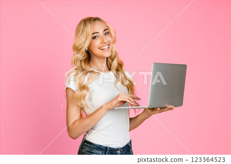 A young woman with long, blonde hair smiles while using a laptop in front of a pink background. She is wearing a white t-shirt and blue jeans. 123364523