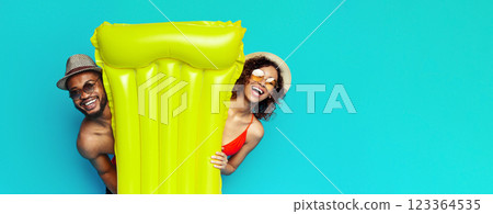 African American happy couple posing together in front of a bright turquoise backdrop, wearing summery attire, including hats and sunglasses, and are holding a large yellow inflatable raft 123364535