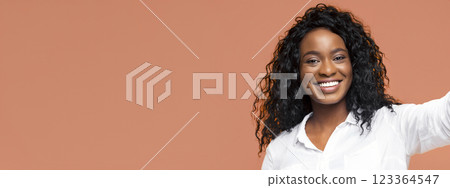 This image features a Black woman with long, curly hair, wearing a white button-down shirt, smiling brightly at the camera. She is posed against a solid peach background. 123364547