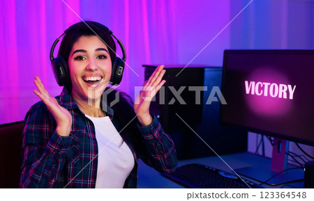 A young woman wearing headphones sits at a computer desk in a dimly lit room, illuminated by pink and blue lights. She is smiling and raising her hands in the air, celebrating her victory. A young woman wearing headphones sits at a computer desk in a dimly lit room, illuminated by pink and blue lights. She is smiling and raising her hands in the air, celebrating her victory. 123364548