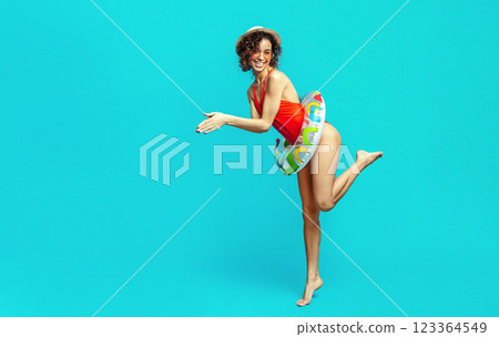 A happy black woman with curly hair is wearing a red swimsuit and a straw hat. She is holding an inflatable ring in one hand, and she is posing against a blue background. 123364549
