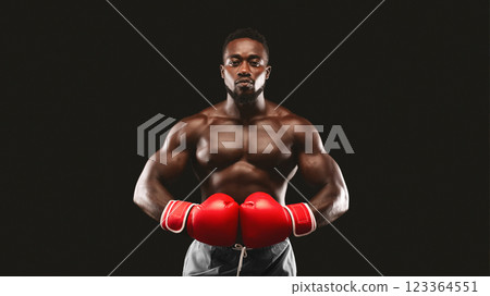 A muscular, shirtless man wearing red boxing gloves stands with his arms raised, ready to fight, against a black background. 123364551