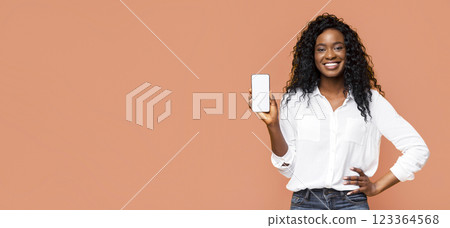 African American young woman with long, curly black hair wearing a white button-down shirt and jeans. She is smiling at the camera while holding up a smartphone with a blank screen in her right hand 123364568