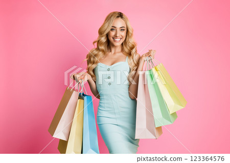 A smiling woman in a light blue dress is standing in front of a pink background, holding multiple colorful shopping bags in her hands. She appears to be happy and satisfied with her purchases. A smiling woman in a light blue dress is standing in front of a pink background, holding multiple colorful shopping bags in her hands. She appears to be happy and satisfied with her purchases. 123364576