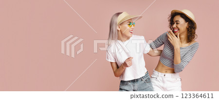 This image shows two women, one with blonde hair and wearing a visor and sunglasses, and the other with dark curly hair, laughing together in front of a pale pink background, copy space 123364611