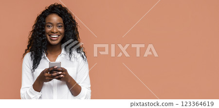 A young Black woman with curly hair smiles while holding a smartphone in her hands. She is dressed in a white button-down shirt and stands in front of a peach background. 123364619