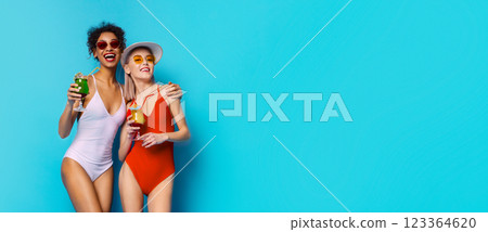 Two women wearing swimsuits and sunglasses are standing in front of a blue background. They are holding cocktails and smiling at each other. Women appear to be enjoying a summer day out. 123364620