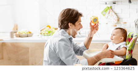 A father sits in a kitchen with a baby in a high chair. He is holding a yellow toy and making the baby laugh. The kitchen is bright and clean. 123364625
