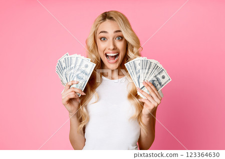 A blonde woman with a white shirt stands in front of a pink background, smiling widely and holding stacks of dollar bills in each hand. She appears to be celebrating a financial success or a win. 123364630