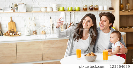 A happy family is taking a selfie in a modern kitchen. The woman is holding a smartphone and smiling at the camera, while the man holds their baby, copy space 123364659