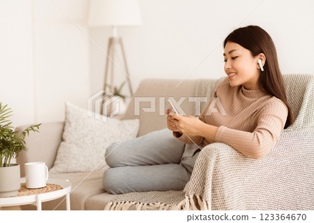 A woman sits comfortably on a couch, wearing earbuds and looking at her phone. A woman sits comfortably on a couch, wearing earbuds and looking at her phone. 123364670