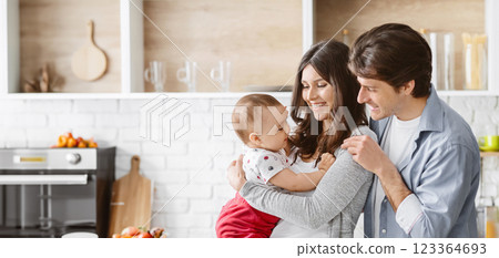 This image shows a happy family of three in their kitchen. The parents are smiling and holding their baby in their arms. The baby is looking up at the parents and appears to be happy. This image shows a happy family of three in their kitchen. The parents are smiling and holding their baby in their arms. The baby is looking up at the parents and appears to be happy. 123364693