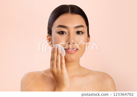 A young Asian woman with dark hair and a light complexion applies makeup to her face with a cotton pad. She is looking directly at the camera and smiling slightly. 123364702
