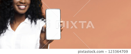 This image shows a smiling black woman holding a smartphone with a blank white screen in front of her. She is wearing a white shirt and standing against a light orange background, cropped 123364709