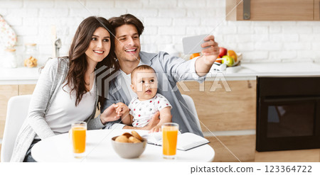 A happy family of three poses for a selfie in their modern kitchen. A young mother sits at the table with her baby, while the father holds the phone and smiles at the camera, copy space 123364722
