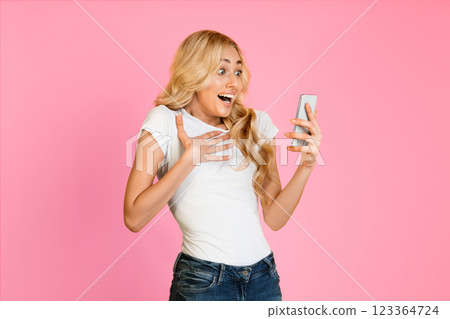 This image shows a surprised woman with blonde hair looking at her phone with her hand on her chest. She is wearing a white t-shirt and blue jeans. The background is a solid pink. This image shows a surprised woman with blonde hair looking at her phone with her hand on her chest. She is wearing a white t-shirt and blue jeans. The background is a solid pink. 123364724