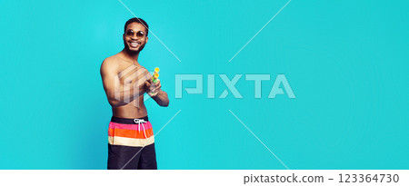 A shirtless black man with a muscular build is wearing colorful swim trunks and sunglasses. He is holding a water gun and smiling at the camera. The background is a solid blue color. 123364730