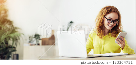 A woman with red hair and glasses smiles while looking at her smartphone. She is sitting at a desk with a laptop and notepad. 123364731