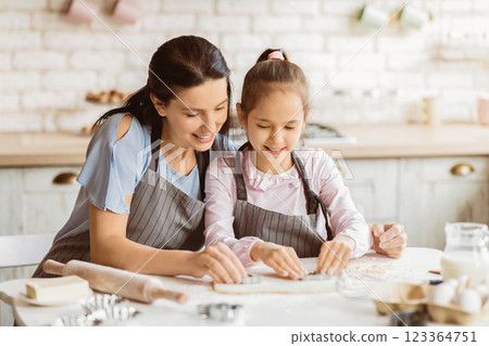 A mother and her young daughter are smiling as they work together to bake cookies in their kitchen. They are both wearing aprons and are focused on their task. The kitchen is clean and bright 123364751