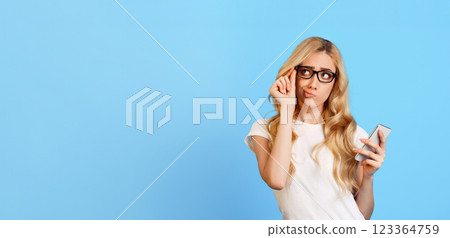 A young woman with blonde hair is holding a smartphone in her right hand while looking away and thinking. She is wearing a white t-shirt and eyeglasses, panorama with copy space A young woman with blonde hair is holding a smartphone in her right hand while looking away and thinking. She is wearing a white t-shirt and eyeglasses, panorama with copy space 123364759